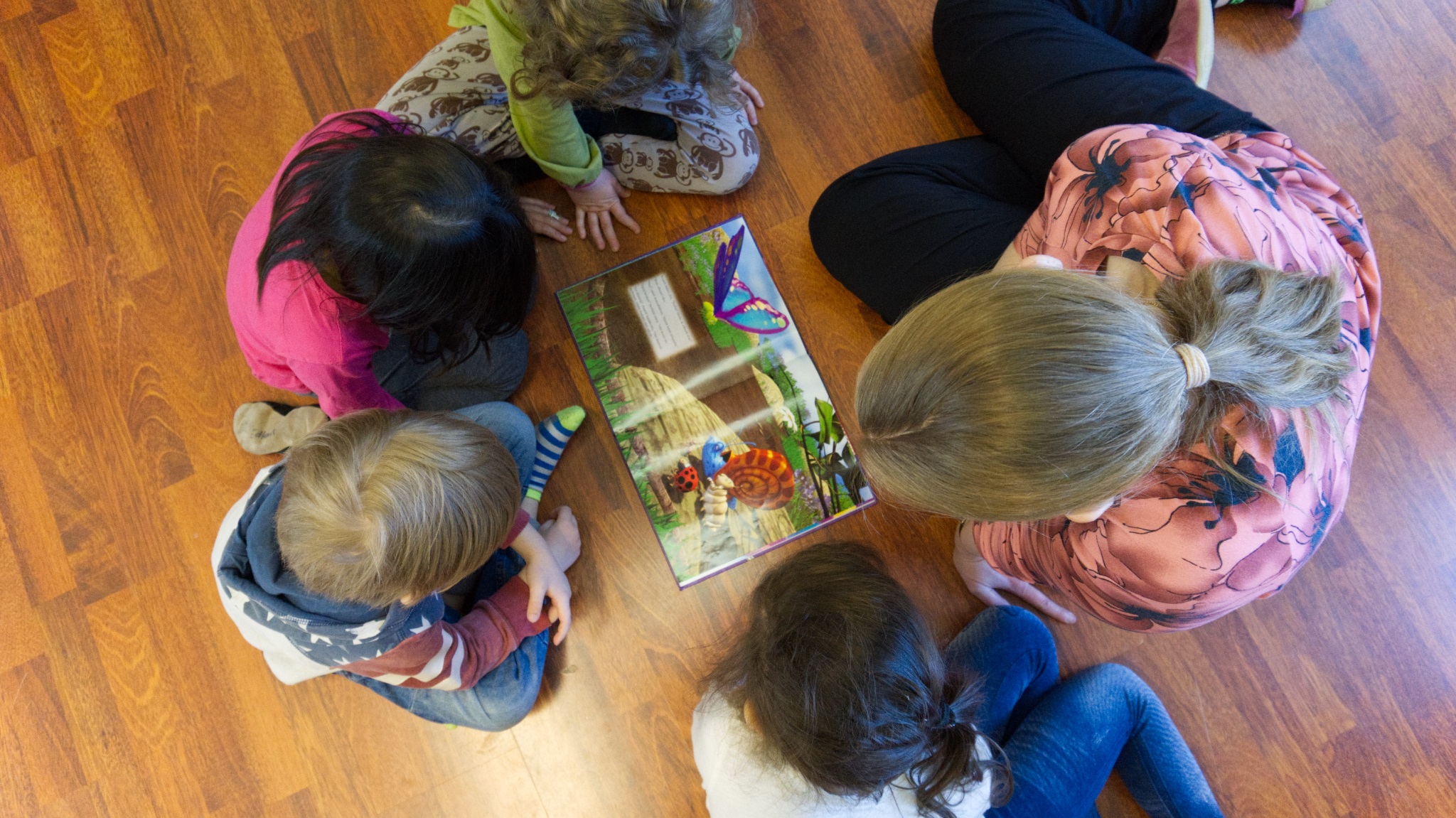 Opettaja ja lapset lattialla ympyrässä lukemassa kirjaa, kuva