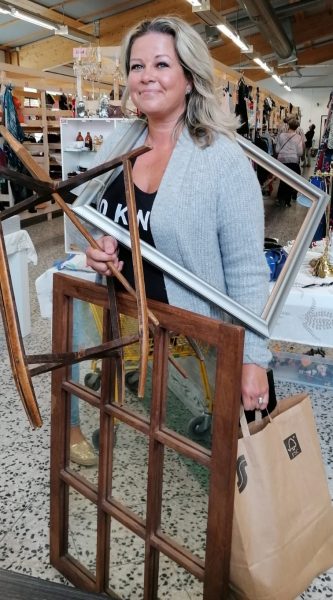 Nainen kirpputorilla kädet täynnä tavaraa ja taulun kehykset kaulassa, kuva